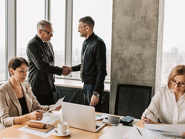 Zwei Frauen arbeiten konzentriert an einem Tisch, auf dem ein Laptop und Diagramme liegen, während sich im Hintergrund zwei Männer die Hand geben