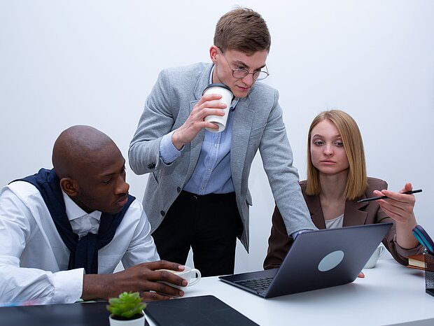 Eine Frau und zwei Männer sitzen am Schreibtisch, auf dem ein Laptop, ein iPad und Ordner liegen, und besprechen ein Thema