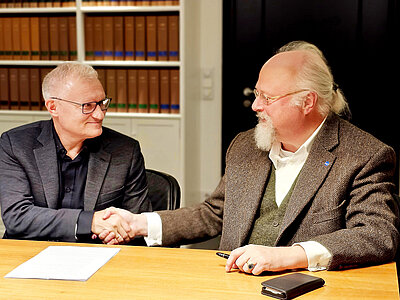 Zwei Männer im Anzug sitzen am Tisch und reichen sich die Hand, auf dem Tisch liegt ein Vertrag und ein Smartphone