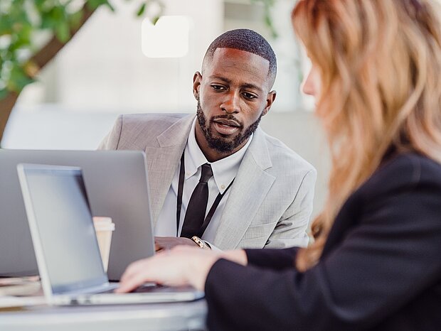 Ein Mann im grauen Anzug und eine Frau sitzen draußen an einem Tisch mit Laptops und besprechen sich
