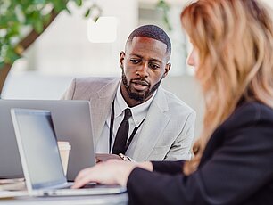 Ein Mann im grauen Anzug und eine Frau sitzen draußen an einem Tisch mit Laptops und besprechen sich