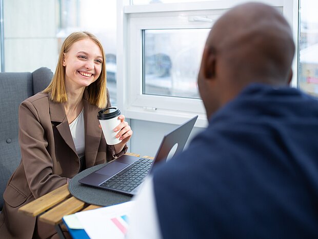Eine lächelnde Frau sitzt mit einem Mann an einem kleinen Tisch, auf dem ein Laptop und Ordner liegen