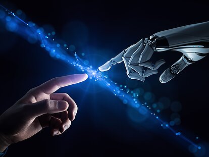 KI-Darstellung, menschlicher und künstlicher Finger treffen sichvor einem dunklen Hintergrund und erzeugen einen schräg von links oben nach rechts unten verlaufenden blau leuchtenden Lichtstrahl.