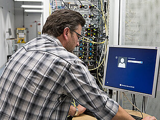Ein Mann im karierten Hemd steht vor dem Serverschrank und meldet sich an seinem PC an