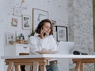 Eine Frau sitzt am Tisch vor dem Laptop und telefoniert. Im Hintergrund sind Bilder zu sehen