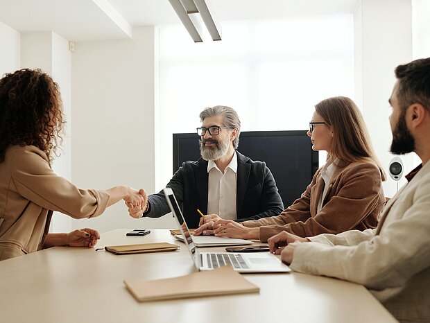 Eine Frau und ein Mann geben sich die Hand, während mehrere Personen an einem Tisch sitzen, auf dem ein Laptop und Schreibblöcke liegen