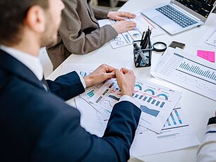 Ein Schreibtisch mit Diagrammen und einem Laptop. Im Vordergrund ein Mann im Anzug der in einem Meeting sitzt