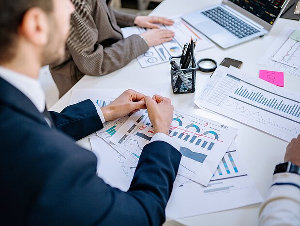 Ein Schreibtisch mit Diagrammen und einem Laptop. Im Vordergrund ein Mann im Anzug der in einem Meeting sitzt