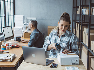 In einem Lager sind zwei Mitarbeiter, ein Mann und eine Frau. Der Mann sitzt vor dem Computer, während die Frau vorne steht und eine Kartonbestellung scannt. Auf dem Tisch des Mannes liegen Tesa, Zahlungsmaschinen 