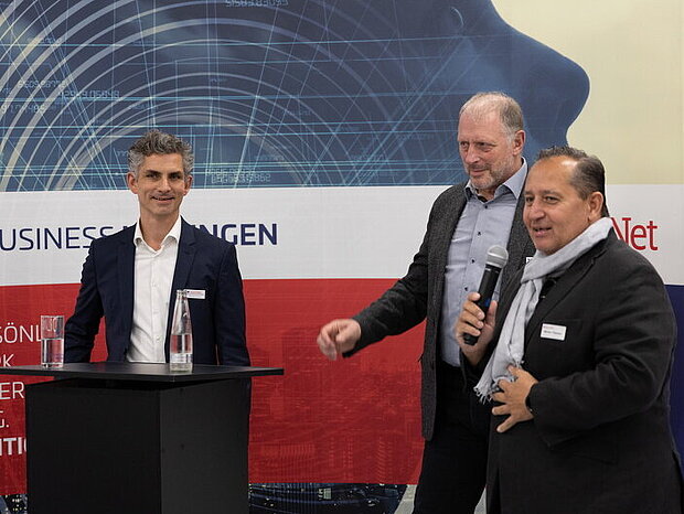 Drei Männer in Anzügen stehen vor einem Rednerpult, einer spricht mit einem Mikrofon in der Hand