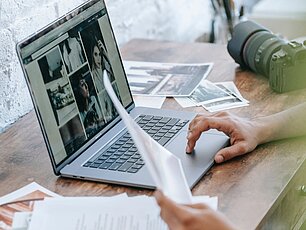 Auf einem Tisch befinden sich ein Laptop , Fotokamera und Papier. Auf dem Laptop-Bildschirm sind Bilder zu sehen. Neben dem Tisch liegen Malstifte. Eine Person hält ein Blatt Papier und berührt das Touchpad des Laptops.