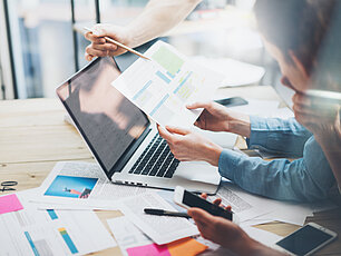 Drei Personen arbeiten zusammen. Auf dem Tisch vorne steht ein Laptop mit vielen Papieren, und eine Frau hält ihre Hände.