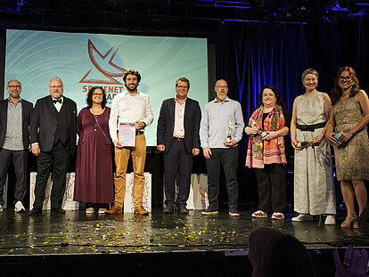 Mehrere Personen stehen nebeneinander auf einer Bühne, die mit goldenem Konfetti bedeckt ist