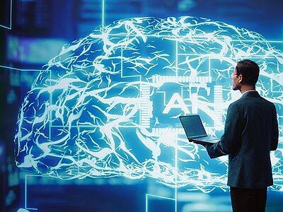 Ein Man mit einem Laptop in der Hand, vor einem digitalem Gehirn. 