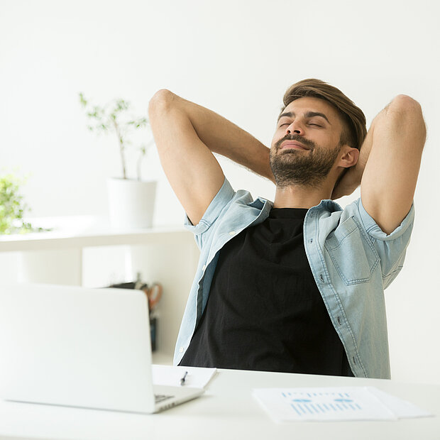 Ein Mann sitzt auf einem Stuhl. Vor ihm steht ein Tisch mit einem Laptop, einer Tasse und Notizzetteln. Der Mann hat die Hände hinter dem Kopf und die Augen geschlossen.