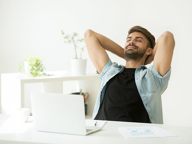 Ein Mann sitzt auf einem Stuhl. Vor ihm steht ein Tisch mit einem Laptop, einer Tasse und Notizzetteln. Der Mann hat die Hände hinter dem Kopf und die Augen geschlossen.