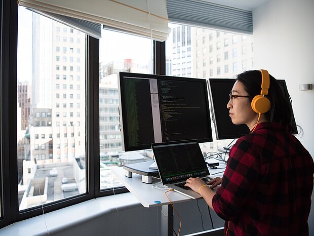 Eine Frau arbeitet an ihrem Schreibtisch mit ihrem Laptop und mehreren Bildschirmen, während sie durch die Glasfenster auf die Stadt blickt