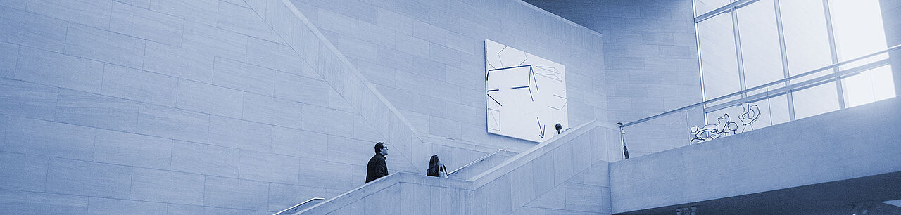 Zwei Personen gehen die Treppen in der Eingangshalle hinauf, im Hintergrund hängen Kunstwerke