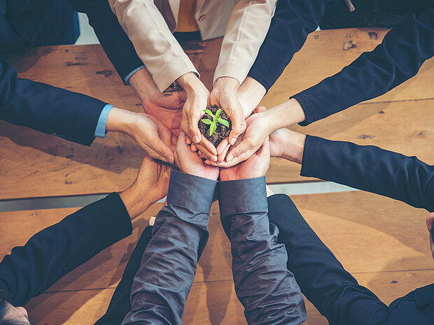 Eine Frau hält eine Pflanze, während mehrere Personen von unten die Hände stützen