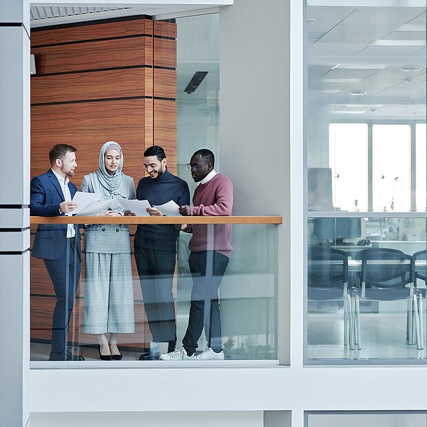 Vier Personen stehen im Büro und halten Blätter in den Händen. Im Hintergrund sind Schreibtische und Bürostühle zu sehen